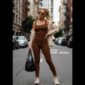 Woman wearing brown Avis tummy control jumpsuit with beige jacket and white sneakers standing on city street